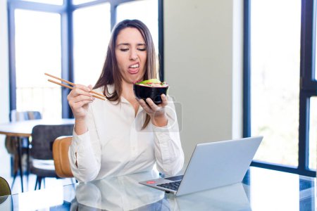 Téléchargez les photos : Jolie jeune adulte femme teleconmmuting avec un ordinateur portable et manger japonais nouilles - en image libre de droit