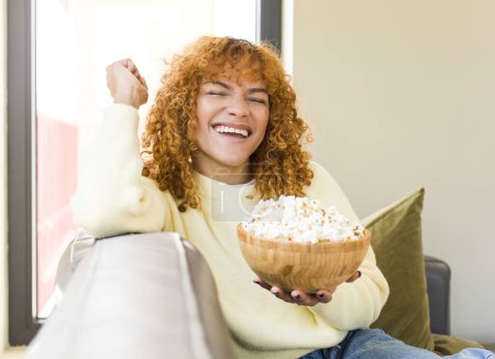 Foto de Young red hair latin pretty woman eating popcorns on a couch and watching a film - Imagen libre de derechos