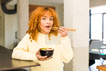 Téléchargez les photos : Jeune roux cheveux latins jolie femme manger ramen nouilles bol à la maison - en image libre de droit