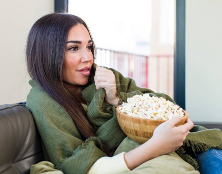 Téléchargez les photos : Jeune jolie femme manger popcorns assis sur un canapé à la maison - en image libre de droit