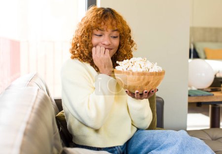 Foto de Joven pelirroja latina bonita mujer comer palomitas de maíz en un sofá y ver una película - Imagen libre de derechos