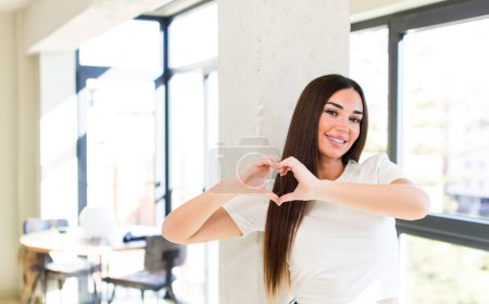 Téléchargez les photos : Young adult pretty woman smiling and feeling happy, cute, romantic and in love, making heart shape with both hands - en image libre de droit