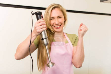 Foto de Young adult pretty blonde chef woman cooking with a hand blender - Imagen libre de derechos