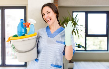Photo for Young pretty woman smiling happily, waving hand, welcoming and greeting you. housekeeper concept - Royalty Free Image
