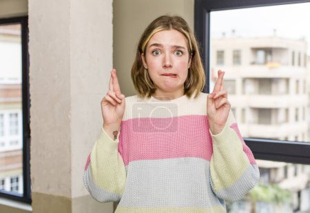 Foto de Pretty caucasian woman crossing fingers anxiously and hoping for good luck with a worried look. home interior concept - Imagen libre de derechos