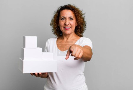 Téléchargez les photos : Pretty middle age woman pointing at camera choosing you. blank white boxes packaging - en image libre de droit