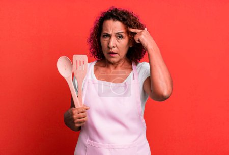 Foto de Pretty middle age woman feeling confused and puzzled, showing you are insane. home wife chef concept - Imagen libre de derechos