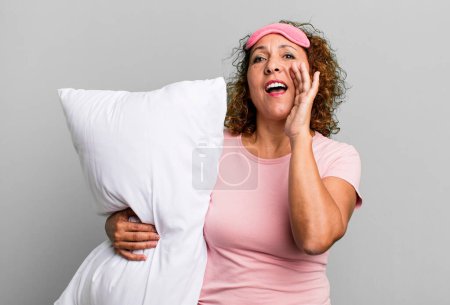 Téléchargez les photos : Pretty middle age woman feeling happy,giving a big shout out with hands next to mouth wearing pajamas night wear and a pillow - en image libre de droit