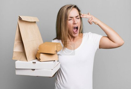 Photo for Pretty blonde woman looking unhappy and stressed, suicide gesture making gun sign. delivery take away food packages concept - Royalty Free Image
