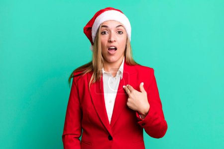 Photo for Pretty blonde woman looking shocked and surprised with mouth wide open, pointing to self. christmas and santa hat concept - Royalty Free Image