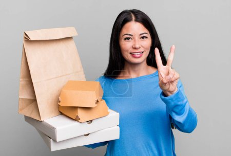 Foto de Hispanic pretty woman smiling and looking friendly, showing number two. with take away fast food packages - Imagen libre de derechos