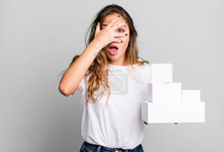 Photo for Hispanic pretty woman looking shocked, scared or terrified, covering face with hand with white boxes packages - Royalty Free Image