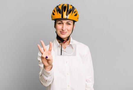 Foto de Joven bonita mujer sonriendo y buscando amigable, mostrando el número tres. concepto de negocio y bicicleta - Imagen libre de derechos