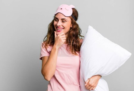 Photo for Hispanic pretty woman smiling with a happy, confident expression with hand on chin wearing pajamas and a pillow - Royalty Free Image