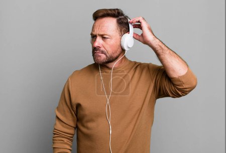 Téléchargez les photos : Homme d'âge moyen se sentant perplexe et confus, tête grattée. écouter de la musique avec un casque - en image libre de droit