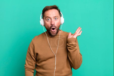 Photo for Middle age man looking surprised and shocked, with jaw dropped holding an object. listening music with a headphones - Royalty Free Image