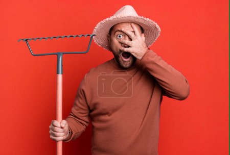 Foto de Middle age man looking shocked, scared or terrified, covering face with hand. farmer with a rake - Imagen libre de derechos