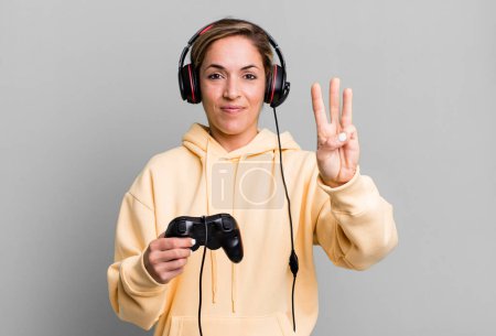 Téléchargez les photos : Pretty blonde woman smiling and looking friendly, showing number three. gamer with headset and a controller - en image libre de droit
