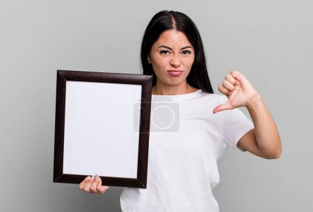 Foto de Hispanic pretty woman feeling cross,showing thumbs down with an empty blank frame - Imagen libre de derechos