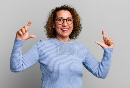 Téléchargez les photos : Femme hispanique d'âge moyen encadrant ou décrivant son propre sourire avec les deux mains, regardant positif et heureux, concept de bien-être - en image libre de droit