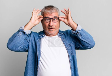 middle age senior man feeling shocked, amazed and surprised, holding glasses with astonished, disbelieving look
