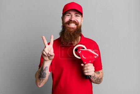 Téléchargez les photos : Barbe longue homme souriant et l'air amical, montrant numéro deux. concept d'emballeur d'expédition - en image libre de droit