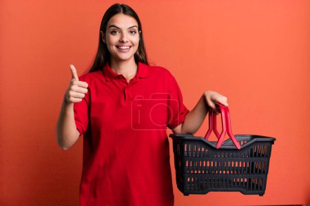 Foto de Young pretty woman feeling proud,smiling positively with thumbs up. empty shopping basket concept - Imagen libre de derechos