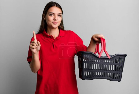 Foto de Young pretty woman smiling and looking friendly, showing number one. empty shopping basket concept - Imagen libre de derechos