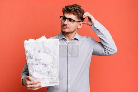 Foto de Young adult caucasian man feeling puzzled and confused, scratching head with a paper balls trash concept - Imagen libre de derechos
