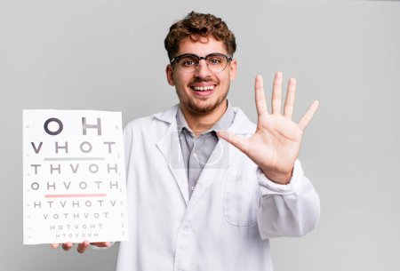 Foto de Young adult caucasian man smiling and looking friendly, showing number five. optical vision test concept - Imagen libre de derechos