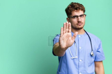 Foto de Young adult caucasian man looking serious showing open palm making stop gesture. nurse concept - Imagen libre de derechos