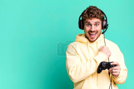 Foto de Young adult caucasian man feeling happy and facing a challenge or celebrating with headset and a controller. gamer concept - Imagen libre de derechos