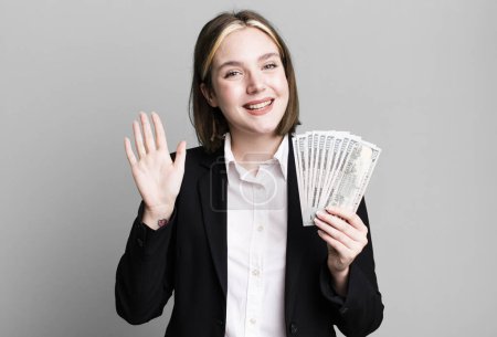 Téléchargez les photos : Young pretty woman smiling happily, waving hand, welcoming and greeting you. dollar banknotes - en image libre de droit