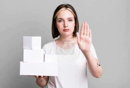 Foto de Joven bonita mujer mirando serio mostrando abierta palma haciendo gesto de parada. cajas blancas en blanco - Imagen libre de derechos