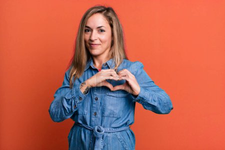 Photo for Blonde adult woman smiling and feeling happy, cute, romantic and in love, making heart shape with both hands - Royalty Free Image