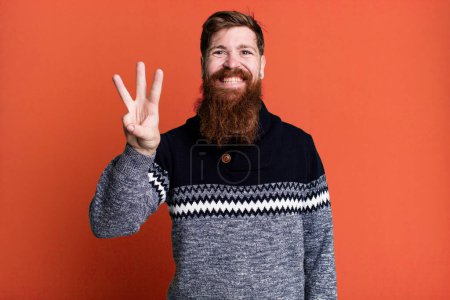 Foto de Barba larga y pelo rojo hombre sonriendo y buscando amigable, mostrando el número tres - Imagen libre de derechos