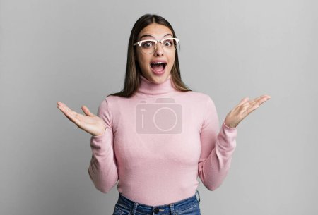 Photo for Pretty young adult woman looking happy and excited, shocked with an unexpected surprise with both hands open next to face - Royalty Free Image