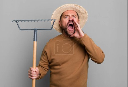 Foto de Hombre de mediana edad sentirse feliz, dando un gran grito con las manos al lado de la boca. granjero con un rastrillo - Imagen libre de derechos