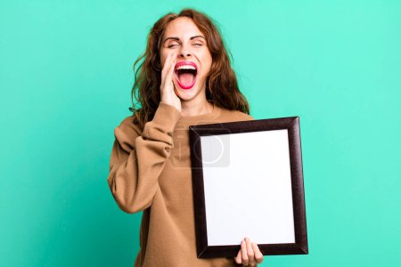 Foto de Hispanic pretty woman feeling happy,giving a big shout out with hands next to mouth with an empty blank frame - Imagen libre de derechos
