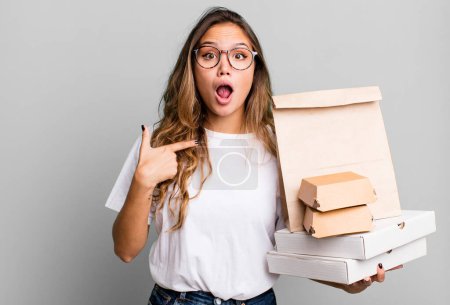 Foto de Hispanic pretty woman looking shocked and surprised with mouth wide open, pointing to self. with fast food packages - Imagen libre de derechos