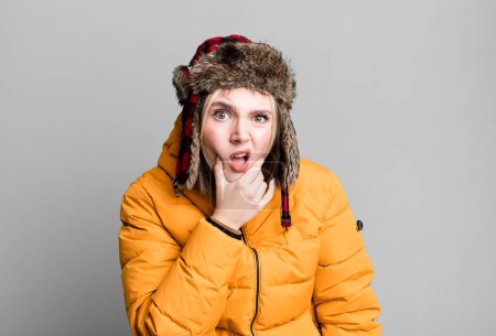 Foto de Young pretty woman with mouth and eyes wide open and hand on chin. cold and coat concept - Imagen libre de derechos