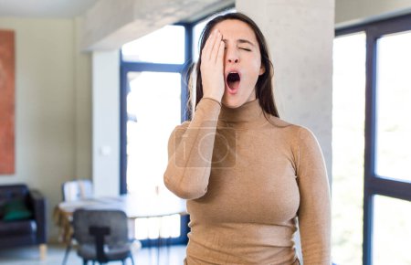 Photo for Young adult pretty woman looking sleepy, bored and yawning, with a headache and one hand covering half the face - Royalty Free Image