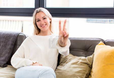 Foto de Young pretty woman smiling and looking happy, carefree and positive, gesturing victory or peace with one hand sitting on a couch - Imagen libre de derechos