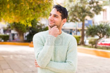 Téléchargez les photos : Jeune homme hispanique souriant joyeusement et rêvant ou doutant, regardant vers le côté - en image libre de droit