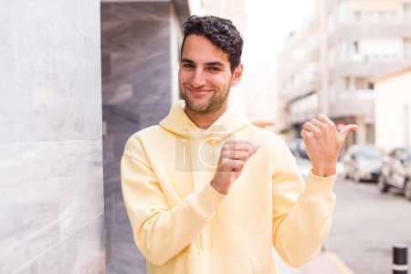 Téléchargez les photos : Jeune homme hispanique souriant joyeusement et avec désinvolture pointant vers copier l'espace sur le côté, se sentant heureux et satisfait - en image libre de droit