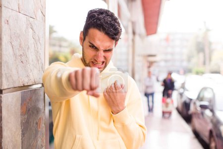 Téléchargez les photos : Jeune homme hispanique regardant confiant, en colère, fort et agressif, avec les poings prêts à se battre en position de boxe - en image libre de droit