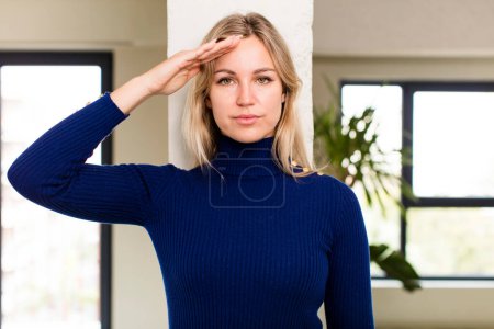 Foto de Joven bonita mujer saludando a la cámara con un saludo militar en un acto de honor y patriotismo, mostrando respeto - Imagen libre de derechos