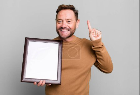 Foto de Hombre de mediana edad sonriendo y buscando amigable, mostrando el número uno con un marco vacío - Imagen libre de derechos