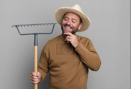 Foto de Hombre de mediana edad sonriendo con una expresión feliz y segura con la mano en la barbilla. granjero con un rastrillo - Imagen libre de derechos
