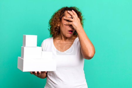 Photo for Pretty middle age woman looking shocked, scared or terrified, covering face with hand. blank white boxes packaging - Royalty Free Image